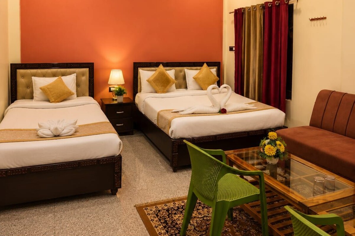 A hotel room with two beds, a green chair, a sofa, a glass coffee table, and a patterned rug.