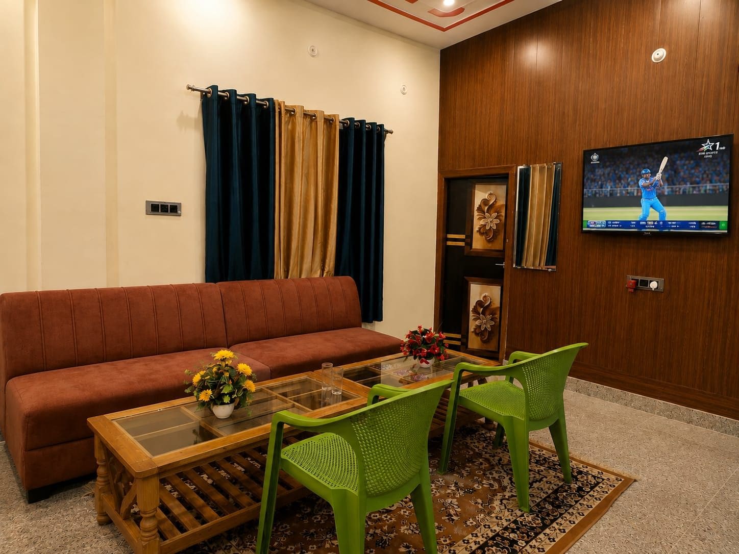 Una sala de estar con un sofá, una mesa de centro con flores, dos sillones verdes, un televisor en la pared y una alfombra estampada.