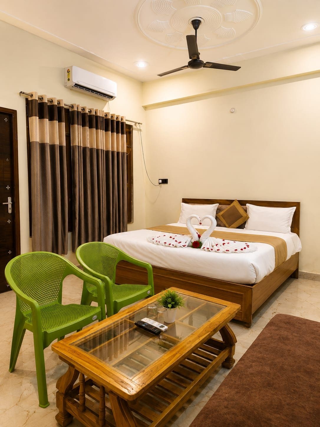 A hotel room with a bed, a ceiling fan, an air conditioning unit, a green chair, a glass-top table, and a brown rug.