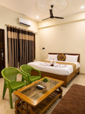 Une chambre d’hôtel avec un lit, un ventilateur de plafond, un climatiseur, un fauteuil vert, une table avec un plateau en verre et un tapis marron.