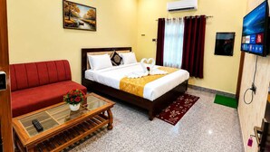 Habitación de hotel con una cama, un sofá rojo, una mesa de centro de vidrio con un jarrón de flores, un equipo de aire acondicionado, un televisor y un cuadro en la pared.