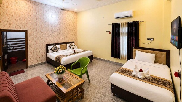 Habitación de hotel con dos camas, un sofá, una silla verde, una mesita con un florero y un equipo de aire acondicionado en la pared.
