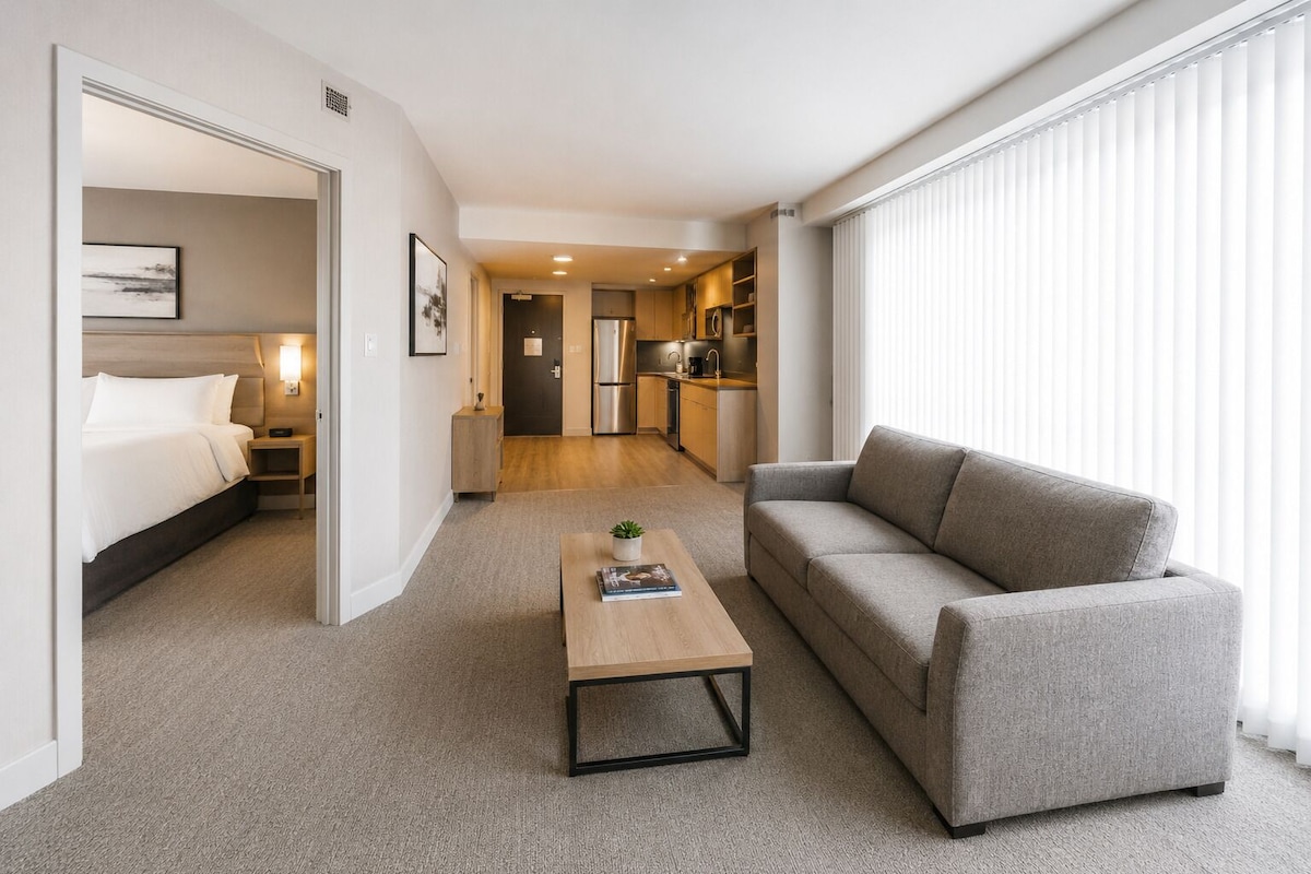 Ein Hotelzimmer mit Bett, Sofa, Couchtisch, einer Küchenzeile und einem großen Fenster mit Jalousien.