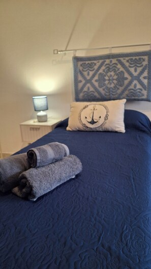 A bed with a blue quilt, a pillow with an anchor design, and rolled towels on the side. - L'Arèna Casa Vacanze elegante vicino al mare (Alghero)