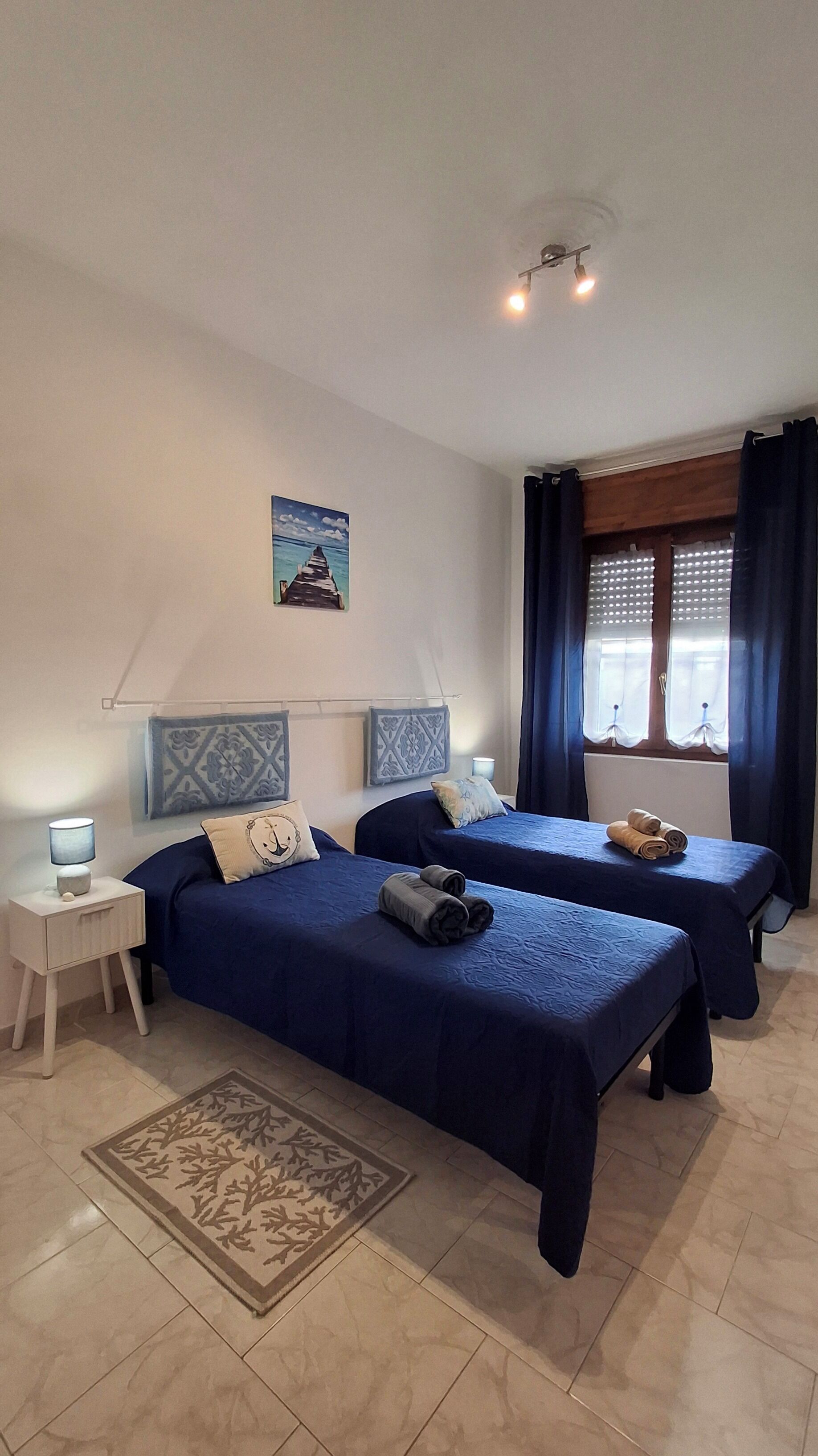 A hotel room with two beds, a nightstand, a lamp, a window with curtains, and a decorative wall piece.