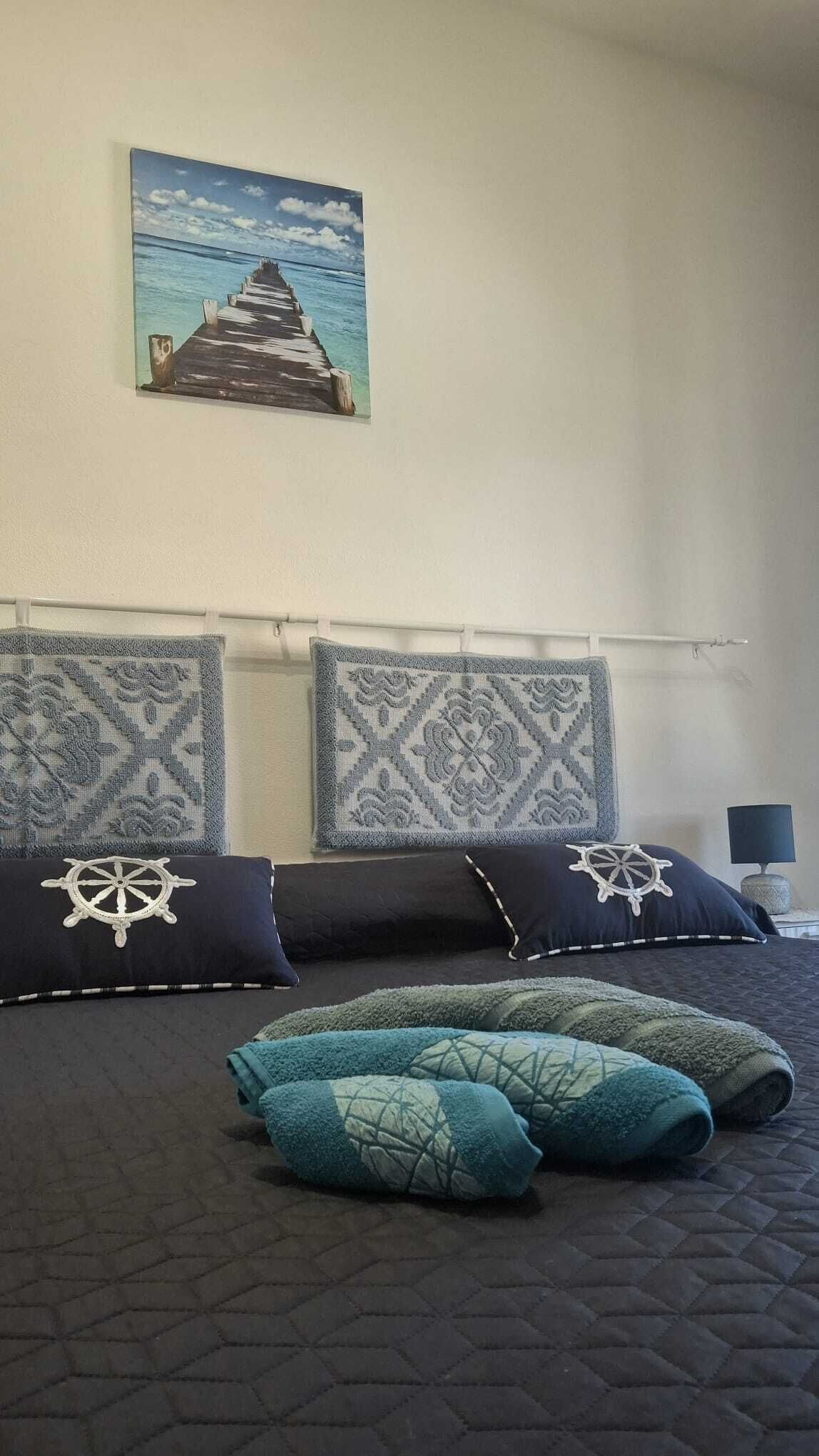 Una cama con un edredón oscuro, dos almohadas con diseño náutico y un cuadro colgado en la pared con una escena costera.