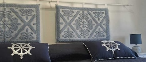 Una cama con un edredón oscuro, dos almohadas con diseño náutico y un cuadro colgado en la pared con una escena costera.
