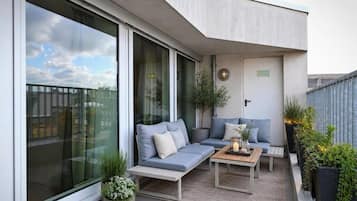Un espacio exterior moderno con zona de estar, un sofá y una mesa, rodeado de plantas en macetas y una barandilla de vidrio.