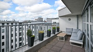 Un balcón con sofá, una mesa, plantas en macetas y vistas de edificios circundantes.