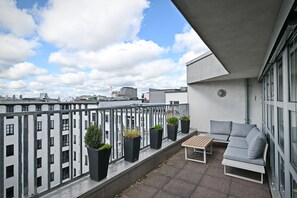 Uma varanda com sofá, mesa, plantas em vasos e vista para edifícios ao redor.
