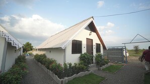 Una casa pequeña con techo inclinado, un porche con barandilla y un jardín con caminos de grava y plantas.