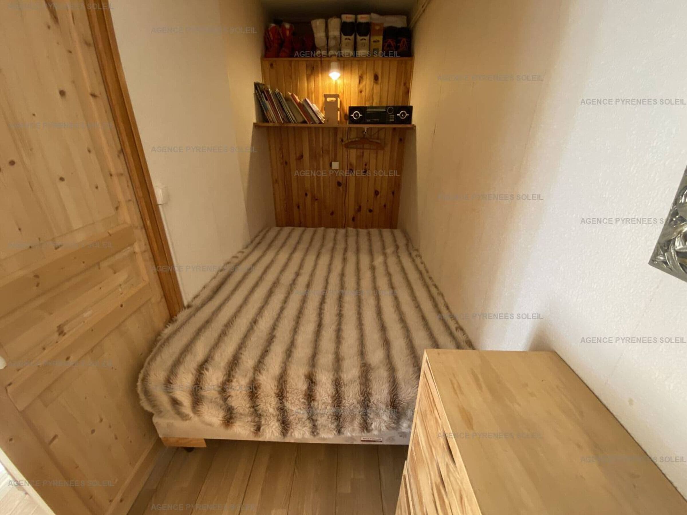 A small, compact room with a bunk bed, a wooden shelf with books and a small lamp, and a wooden bench.