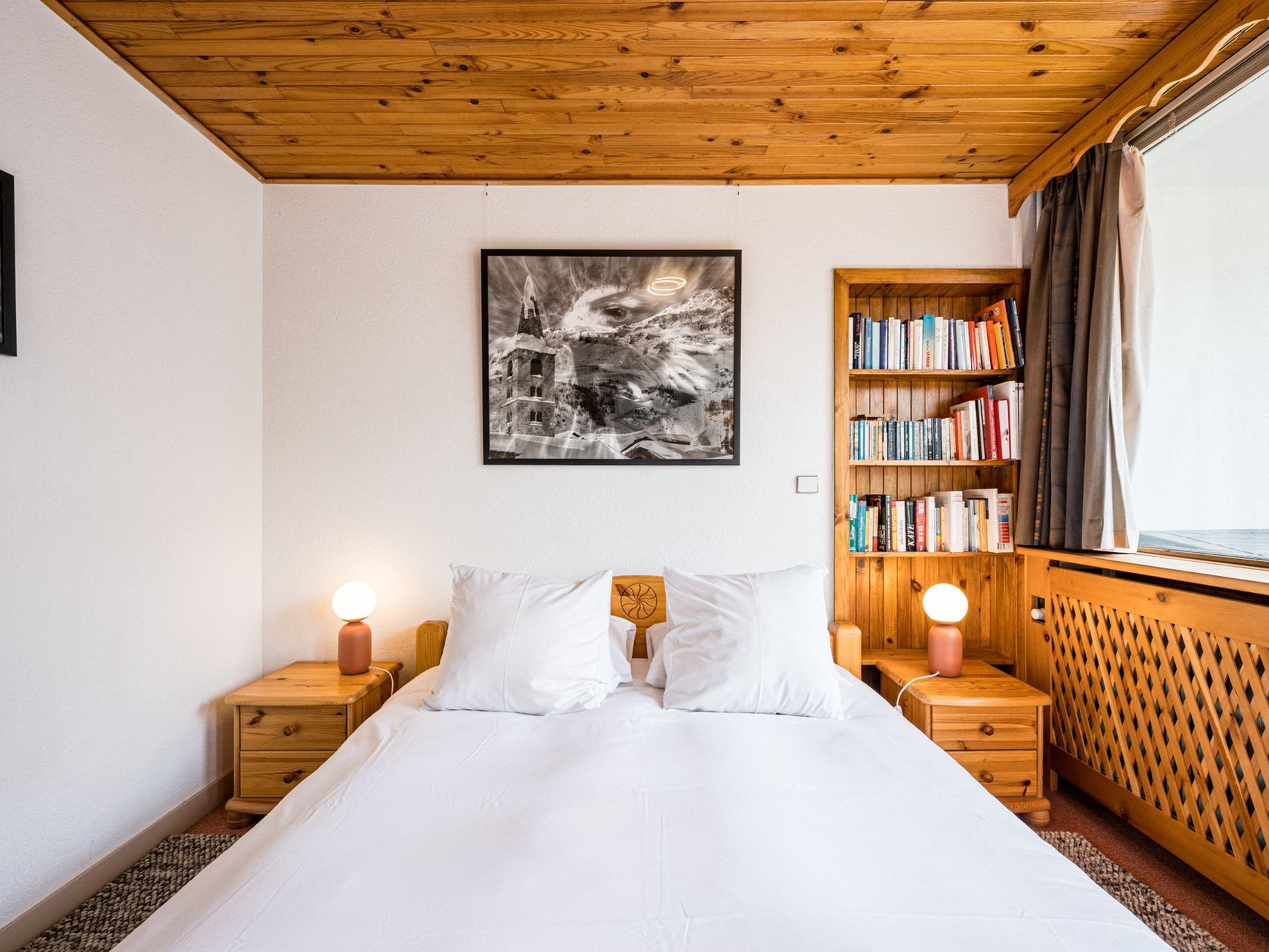 Un dormitorio con techo de madera, una cama con ropa de cama blanca, una estantería y un cuadro enmarcado en la pared.