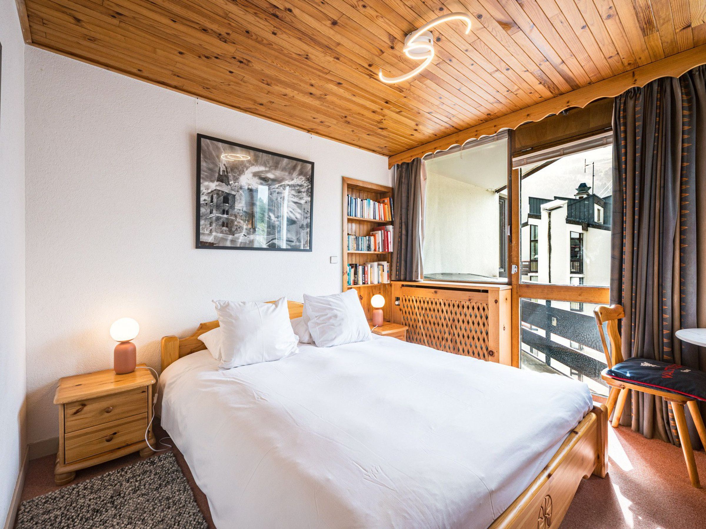A bedroom with a wooden ceiling, a bed with white bedding, a nightstand with a lamp, a bookshelf, and a window with curtains.