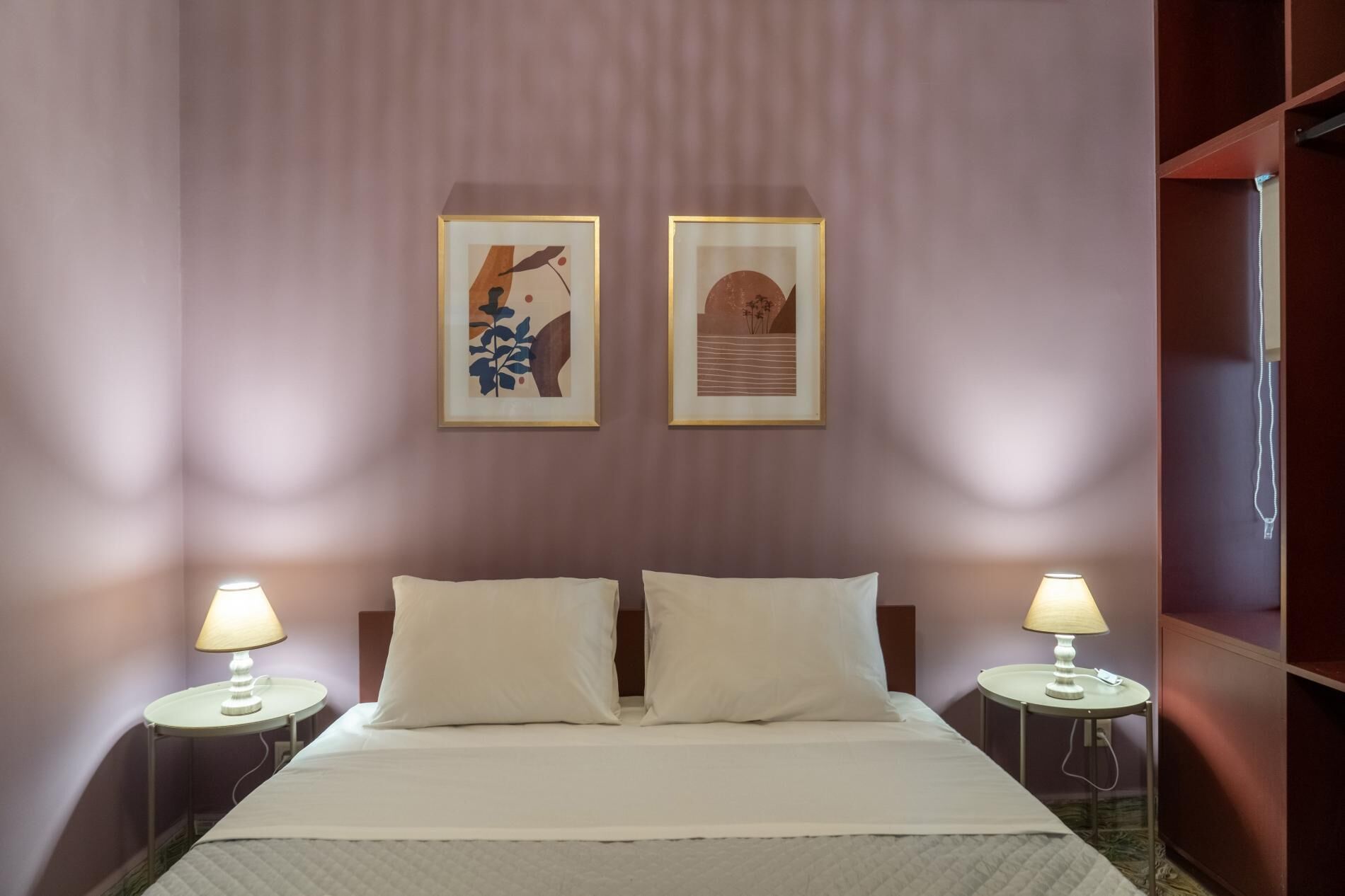 A hotel room with a bed, two bedside lamps, and two framed artworks on the wall.