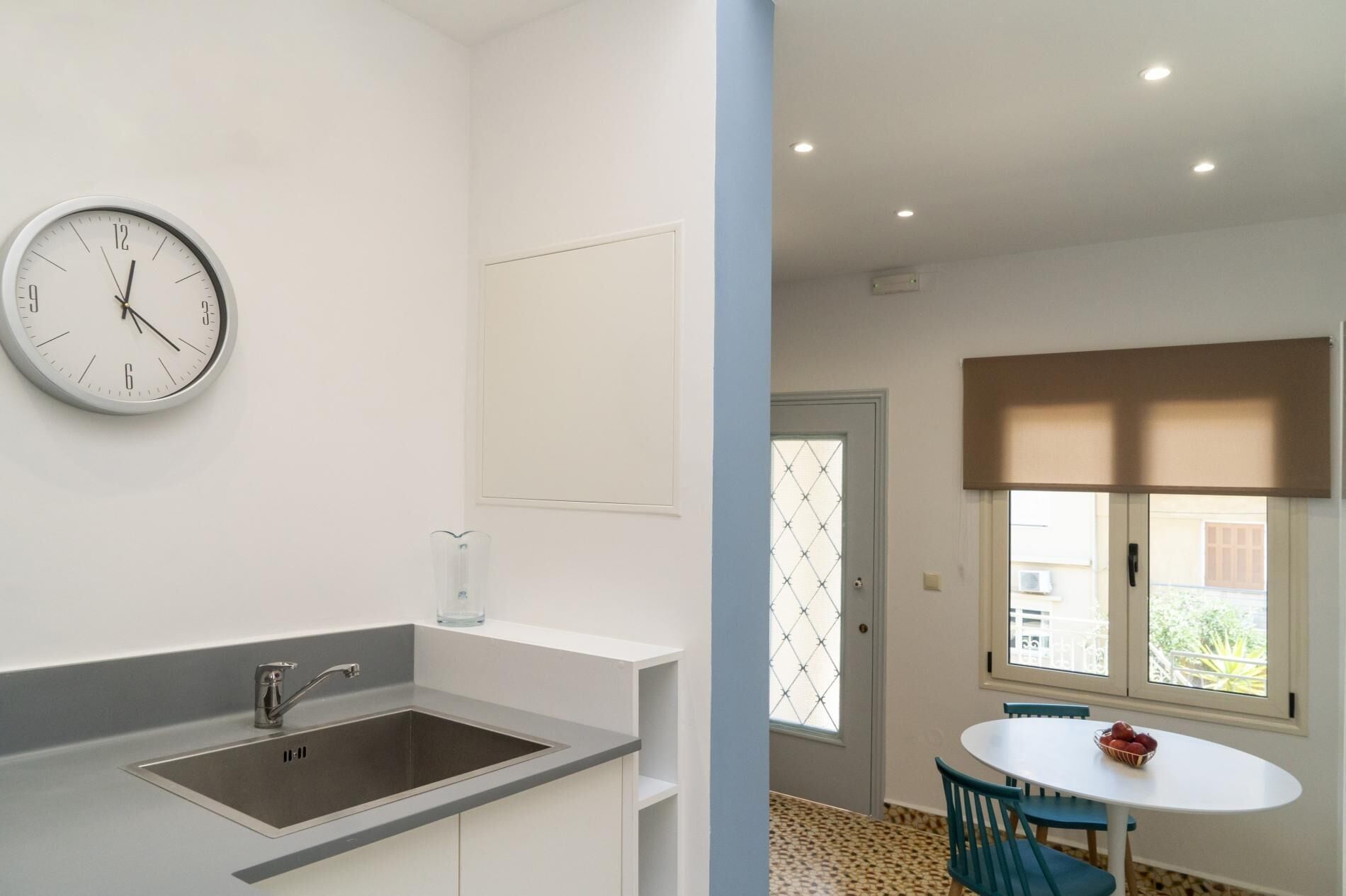 A modern kitchen with a sink and a clock, a dining area with a table and chairs, and a window with a view.