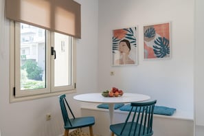 Une pièce comprenant une table ronde, deux chaises, une fenêtre donnant sur des immeubles et deux œuvres d’art accrochées au mur.