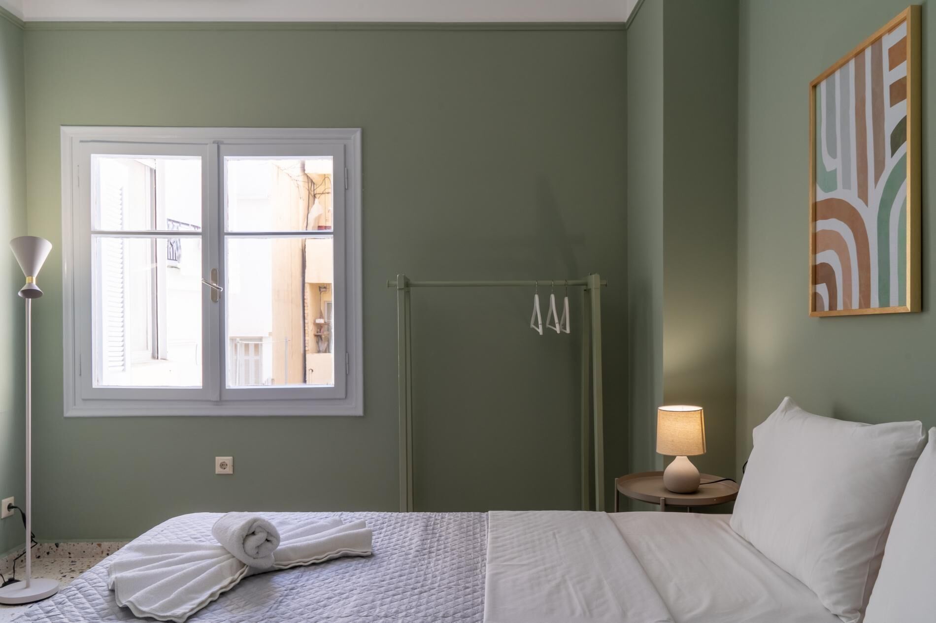 A hotel room with a bed, a window, a door, a shower, a bedside lamp, and a painting on the wall.