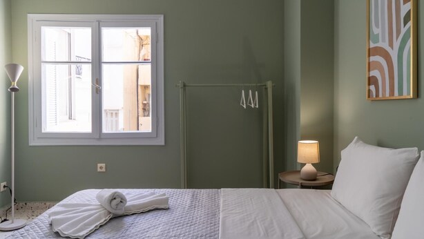 A hotel room with a bed, a window, a door, a shower, a bedside lamp, and a painting on the wall.