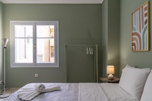 Habitación de hotel con cama, ventana, puerta, ducha, lámpara de noche y un cuadro en la pared.