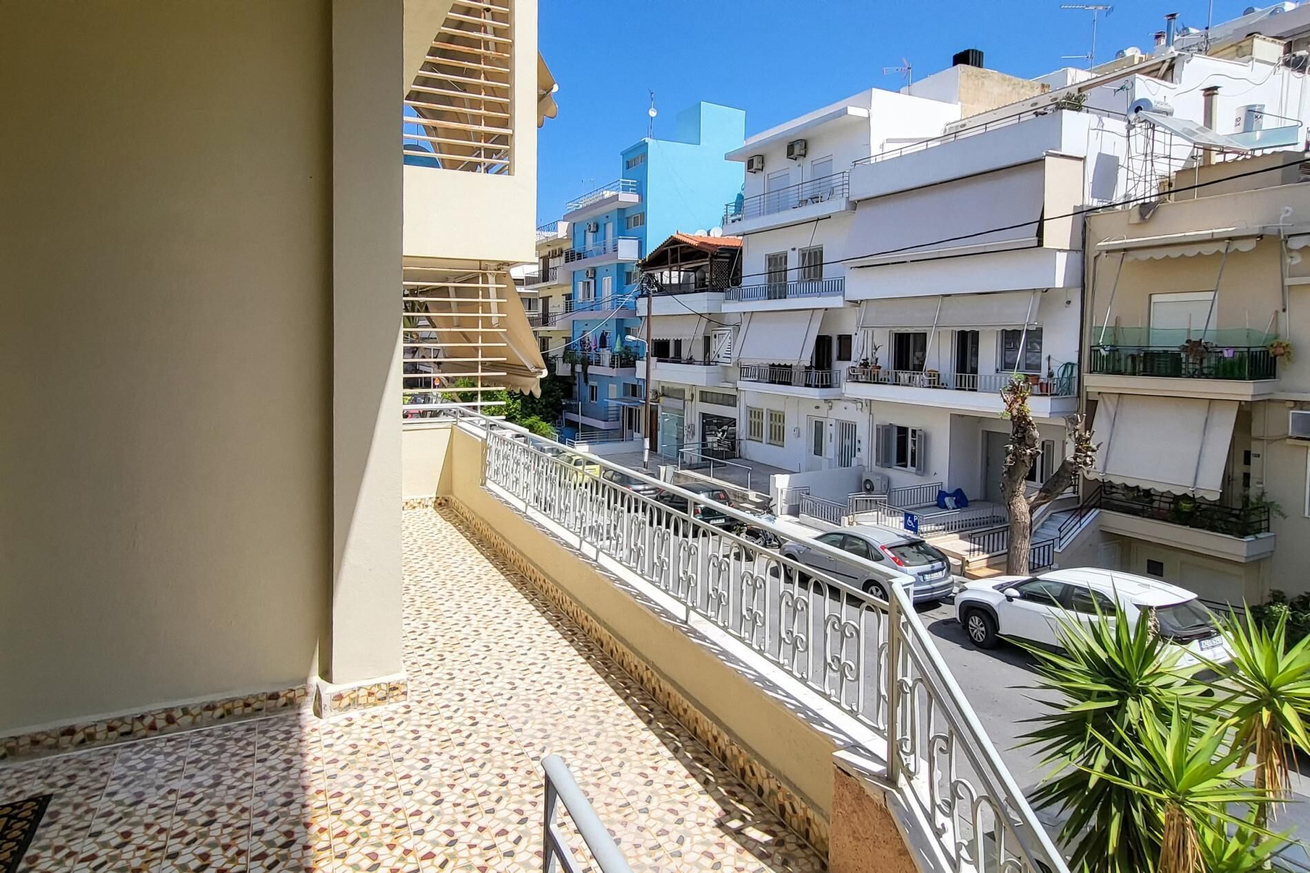 A balcony with a tiled floor, a metal railing, and a view of multi-story buildings with balconies and parked cars.
