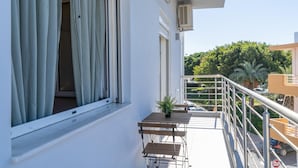 En balkon med gelænder, et lille bord med en potteplante, samt udsigt til et træ og parkerede biler.