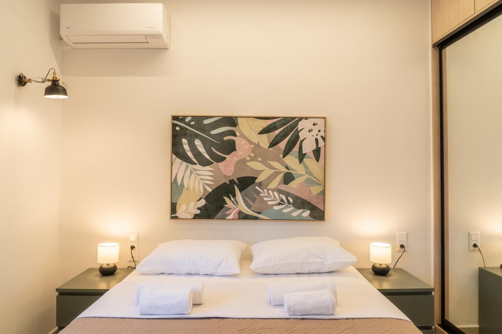 A hotel room with a bed, two bedside tables, a wall-mounted air conditioner, and a floral artwork.