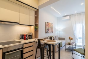 Uma cozinha moderna com forno embutido e cafeteira, uma sala de jantar com mesa e cadeiras, e uma sala de estar com sofá e um quadro.