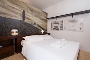 Habitació d'hotel amb un llit, una nitòria amb una làmpada, una paret amb un quadre gran i una estant amb diversos objectes.