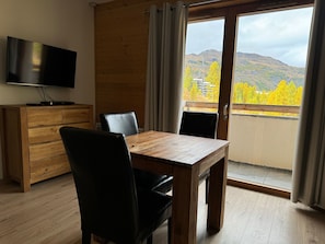Ein Zimmer mit einem Holztisch und Stühlen, einem Fernseher auf einer Holzkommode und Blick auf eine gebirgige Landschaft durch eine Glastür.