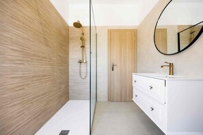 Ein modernes Badezimmer mit einer Holzwand, einer Duschkabine aus Glas, einem weißen Waschtisch mit goldenen Armaturen und einem runden Spiegel.