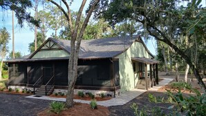 A rustic cabin with a stone foundation, wooden siding, and a gabled roof, surrounded by trees and a gravel driveway.