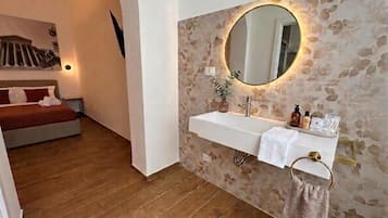 Ein Badezimmer mit Steinwand, einem runden Spiegel, einem weißen Waschbecken und einem Bett im angrenzenden Raum.