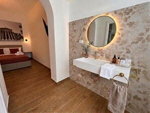 Ein Badezimmer mit Steinwand, einem runden Spiegel, einem weißen Waschbecken und einem Bett im angrenzenden Raum.