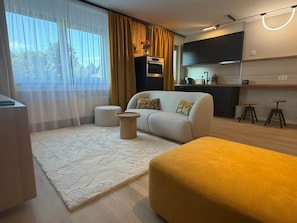 A modern living room with a sofa, ottoman, and a coffee table. There is a kitchen area with a microwave and cabinets.