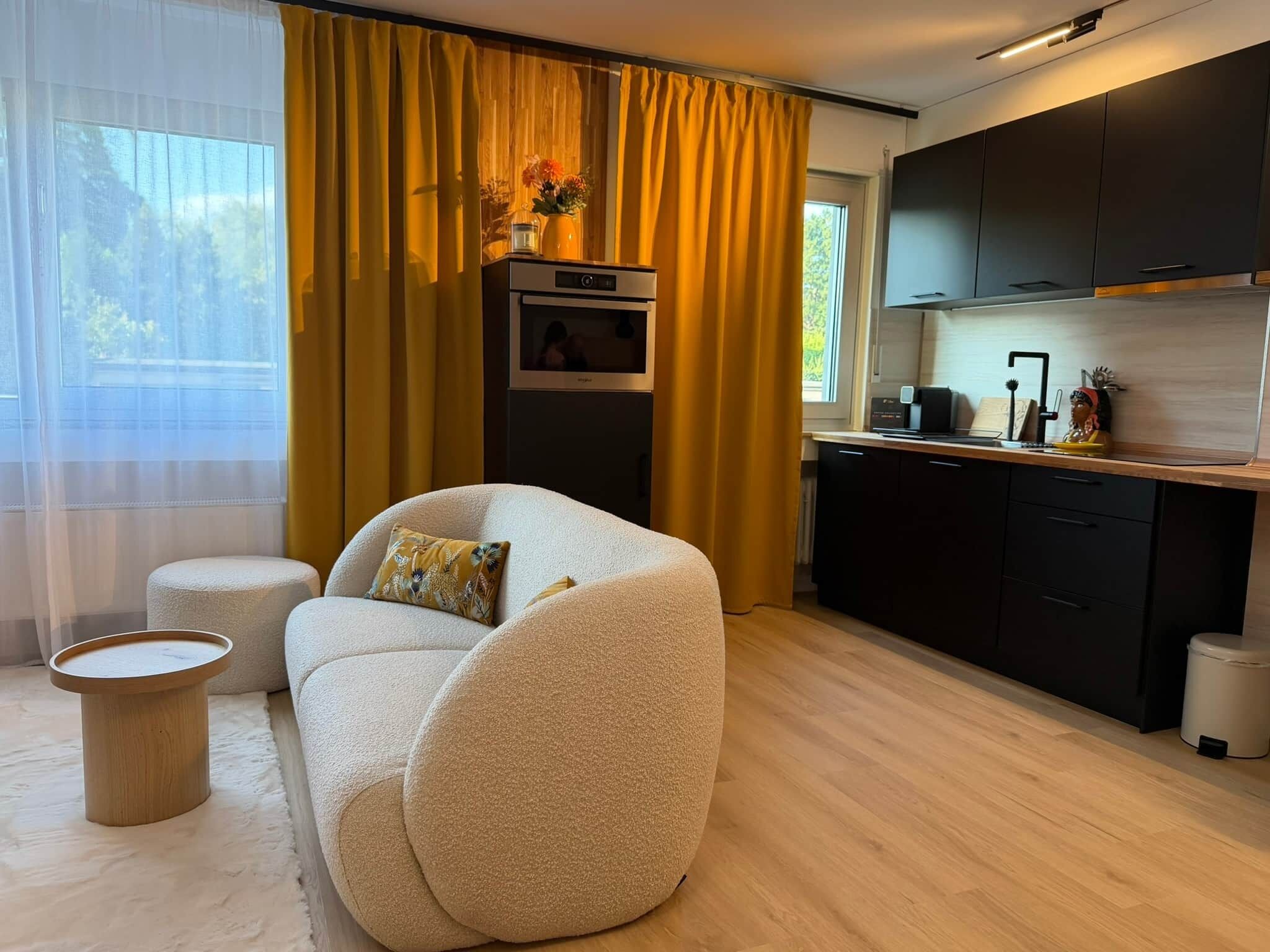 Een moderne woonkamer met een bank, een klein rond tafeltje en een keuken met een koelkast en kastjes.