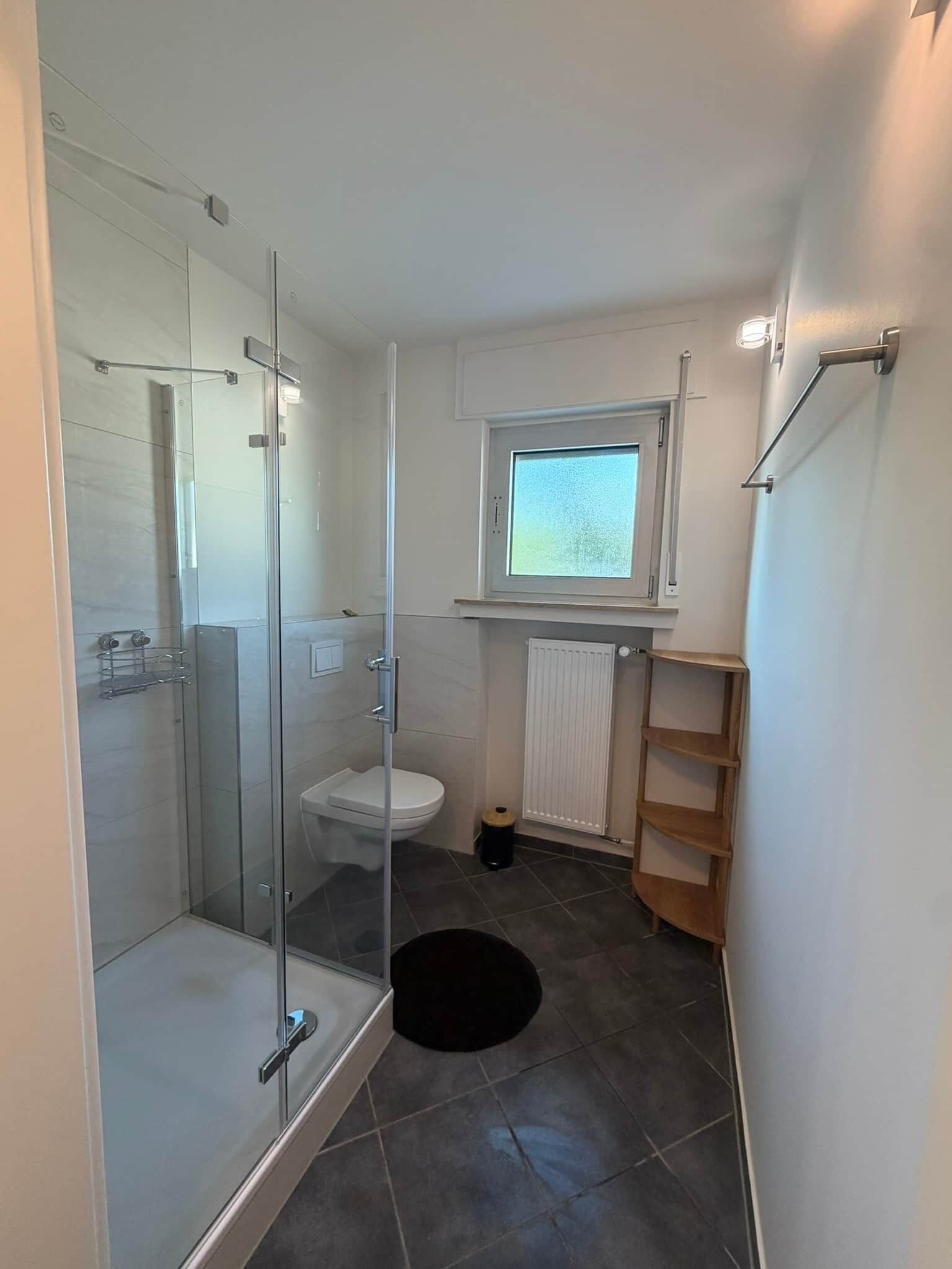 Ein modernes Badezimmer mit einer gläsernen Duschkabine, Toilette und einem Fenster über dem Waschbecken.
