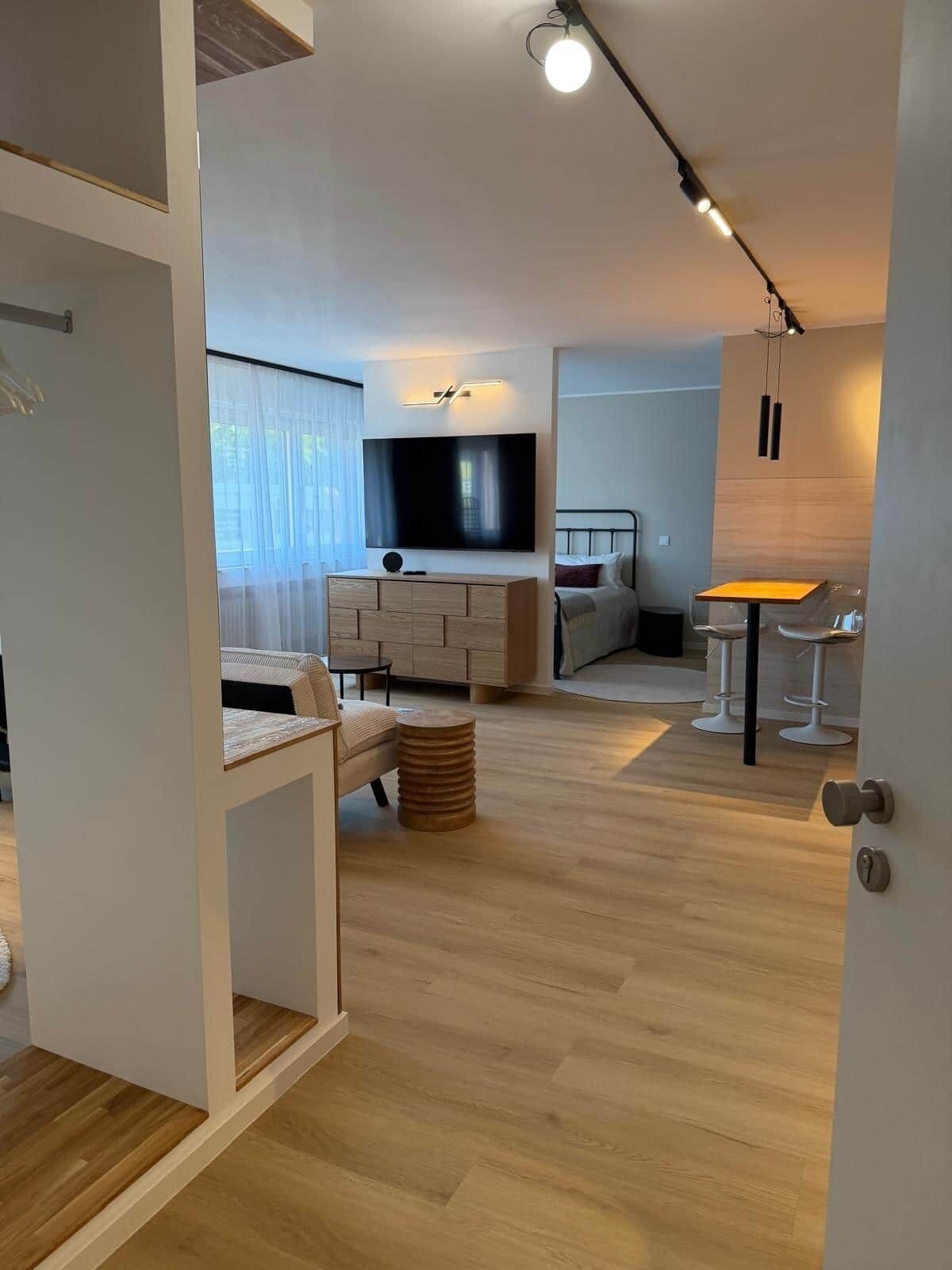 A modern, well-lit room with a wooden floor, a bed, a TV mounted on the wall, and a small table with chairs.