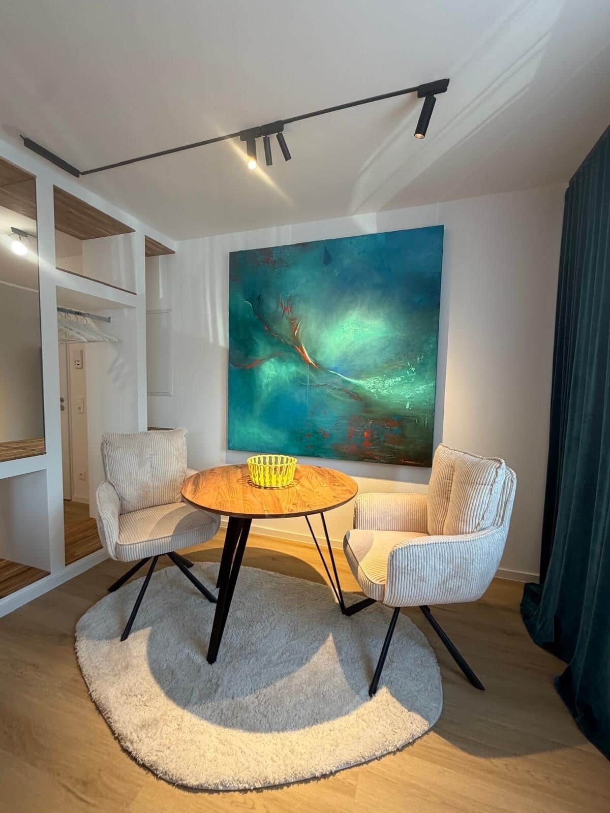 Ein modernes Wohnzimmer mit einem runden Holztisch, zwei gestreiften Sesseln, einem großen abstrakten Gemälde und einer Deckenleuchtenanlage.