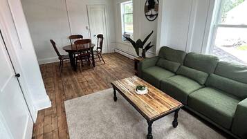A living room with a green sofa, a wooden coffee table, a dining table with chairs, and a view of the outdoors through a window.