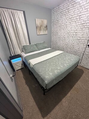 Habitación con una cama individual, colcha de cama gris con estampado, pared blanca con diseño de ladrillo, un cuadro y una cortina.