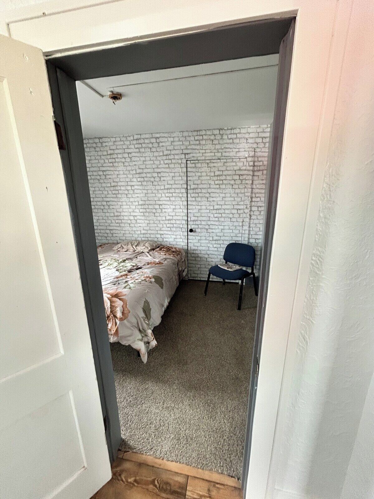 A bedroom with a bed, a chair, a white brick wall, and a door.