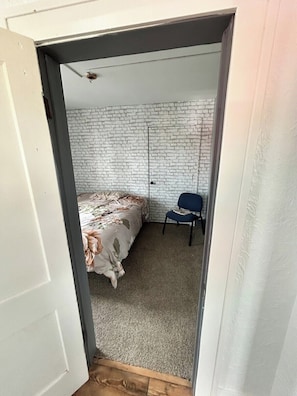 A bedroom with a bed, a chair, a white brick wall, and a door.
