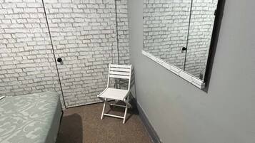 A small, narrow room with a brick-patterned wall, a single bed, a white chair, and a large mirror.