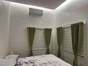A bedroom with a bed, two windows with curtains, a ceiling fan, and a wall-mounted air conditioner.