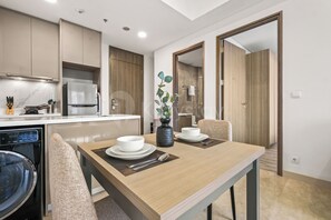 A modern kitchen with a built-in oven, microwave, and dishwasher. There is a dining area with a table set for two, a vase with a plant, and a view of the living room through an open door. - Weska by Kozystay - Cilandak (South Jakarta)