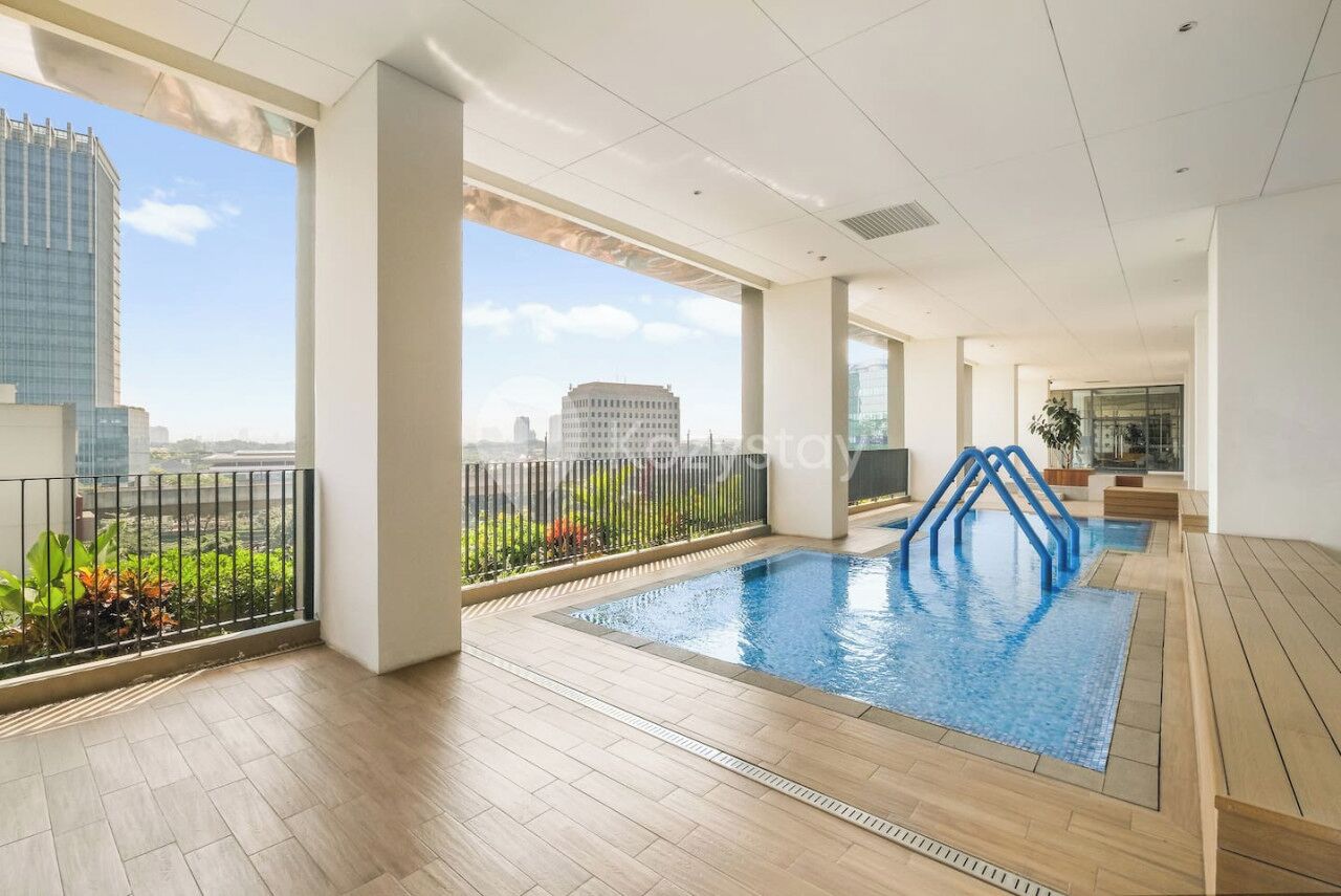 An indoor swimming pool with a view of the cityscape, surrounded by a balcony with a railing and potted plants.