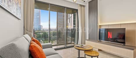 A modern living room with a grey sofa, orange cushions, a round wooden side table with a book, a flat-screen TV, and a city view through large windows.