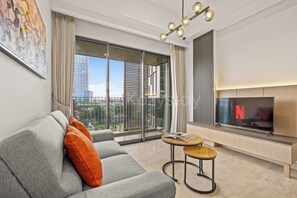 A modern living room with a grey sofa, orange cushions, a round wooden side table with a book, a flat-screen TV, and a city view through large windows. - Weska by Kozystay - Cilandak (South Jakarta)