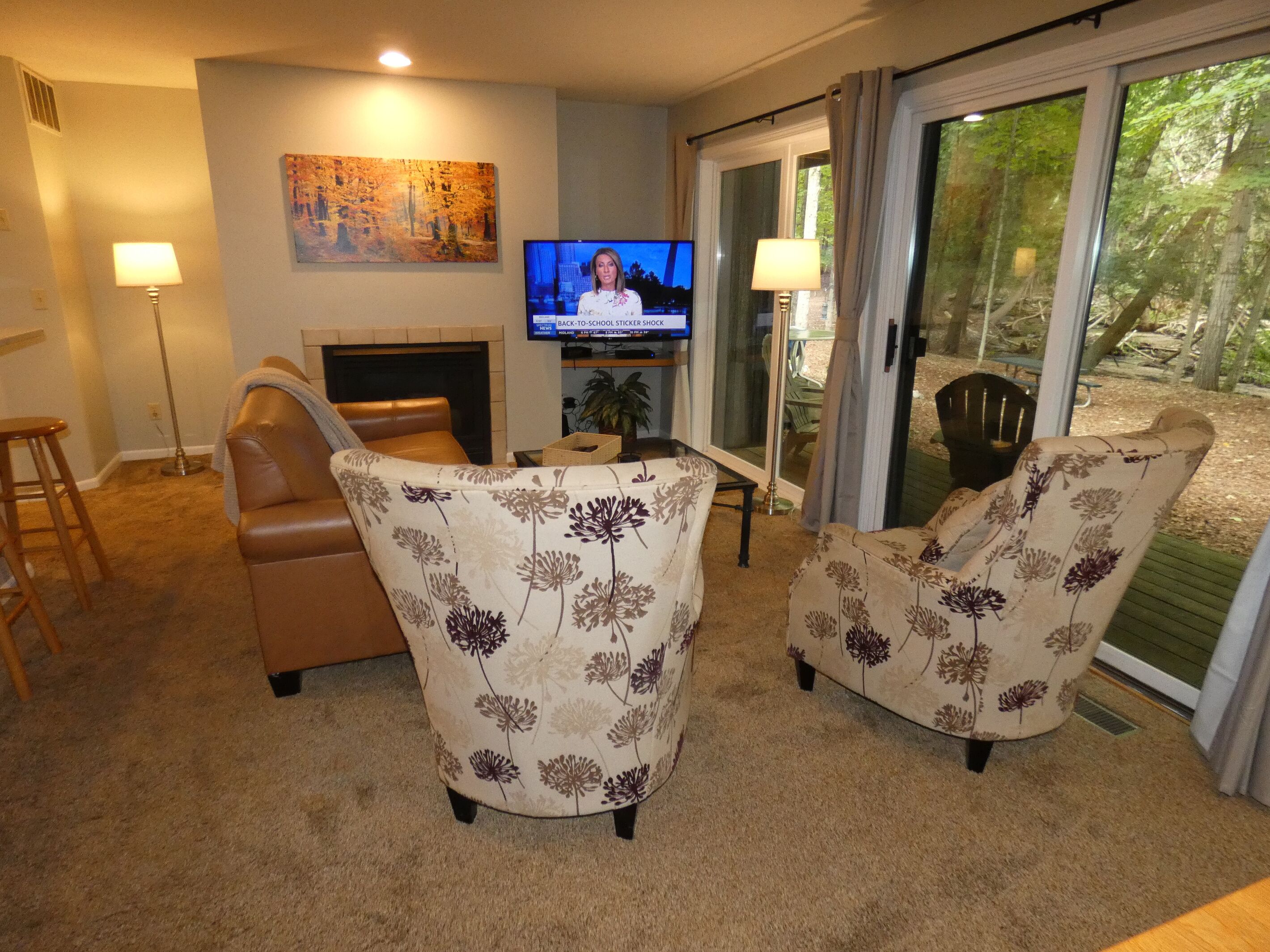 A living room with a fireplace, two floral armchairs, a television, and a sliding glass door leading to an outdoor area.